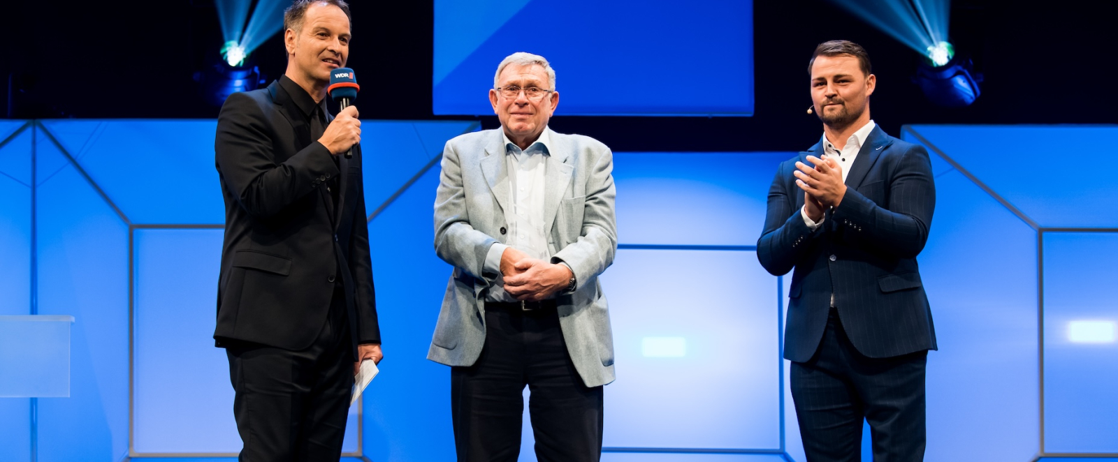 Drei Männer stehen auf einer Bühne, der ältere Herr in der Mitte hat ein hellgraues Sakko an, die beiden anderen Herren tragen dunkelfarbige Anzüge. Der Hintergrund ist bläulich beleuchtet.