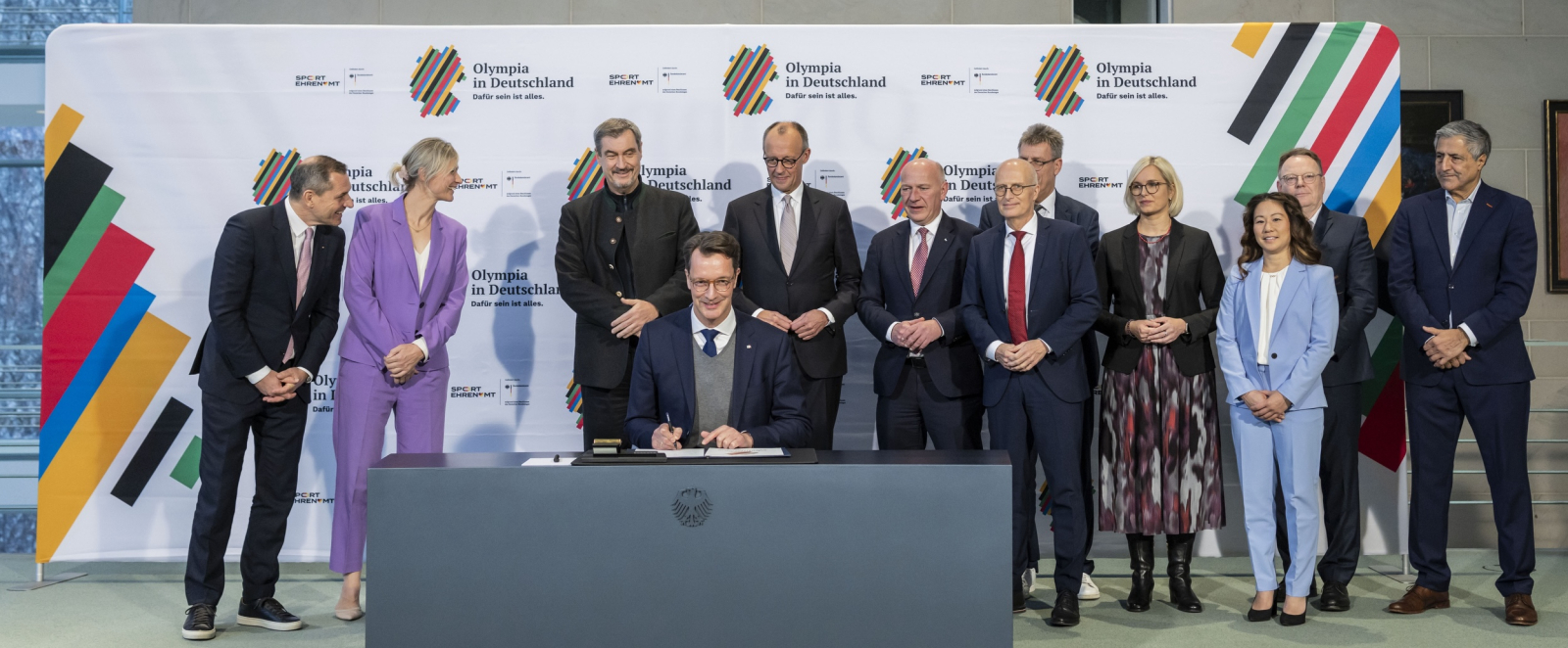 Henrik Wüst an einem Schreibtisch hinter ihm die anderen Akteure bei der Unterzeichnung im Bundeskanzleramt