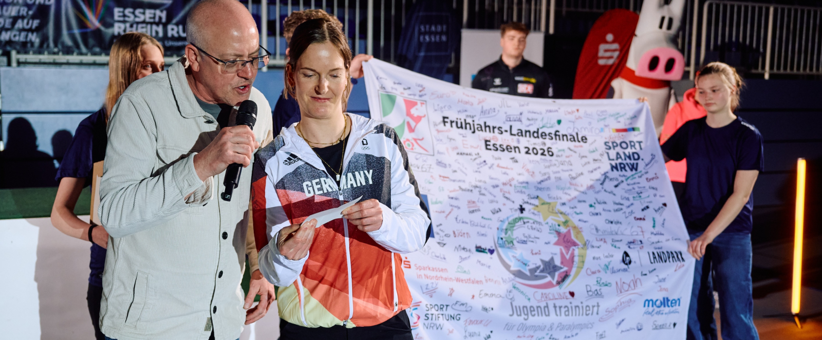 Interviewsituation mit zwei Personen dahinter halten zwei Mädchen ein Banner