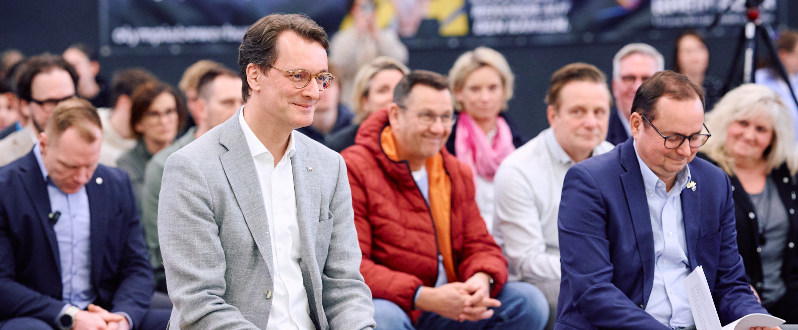 Ministerpräsident Wüst in einem hellen Jacket, in einer Halle mit mehreren Personen, die um ihn herum sitzen