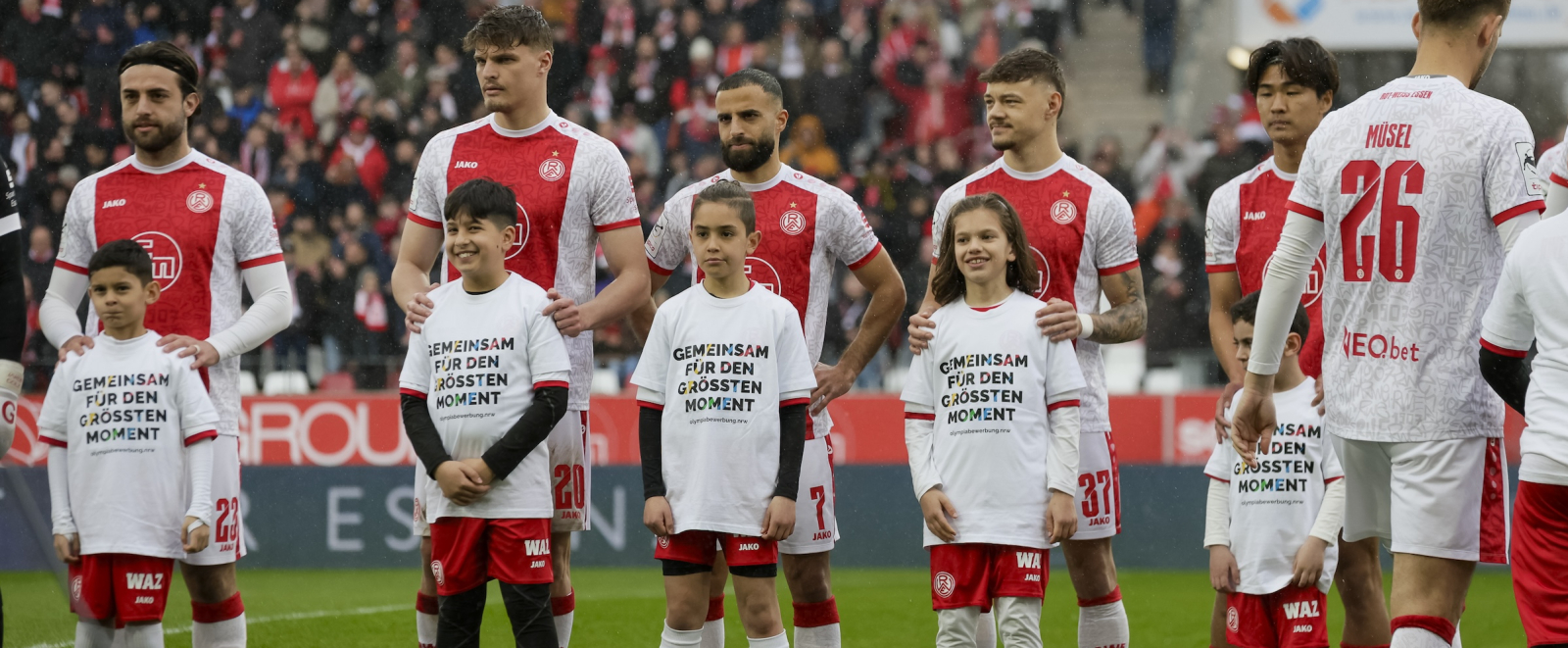 Fußballspieler und Einlaufkinder mit T-Shirts mit der Aufschrift "Gemeinsam für den größten Moment" auf dem Rasen in einem Stadion
