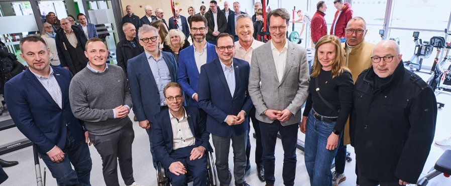 Ministerpräsident Wüst inmitten von Personen, davon eine im Rollstuhl, in einem Raum mit Fenstern im Hintergrund