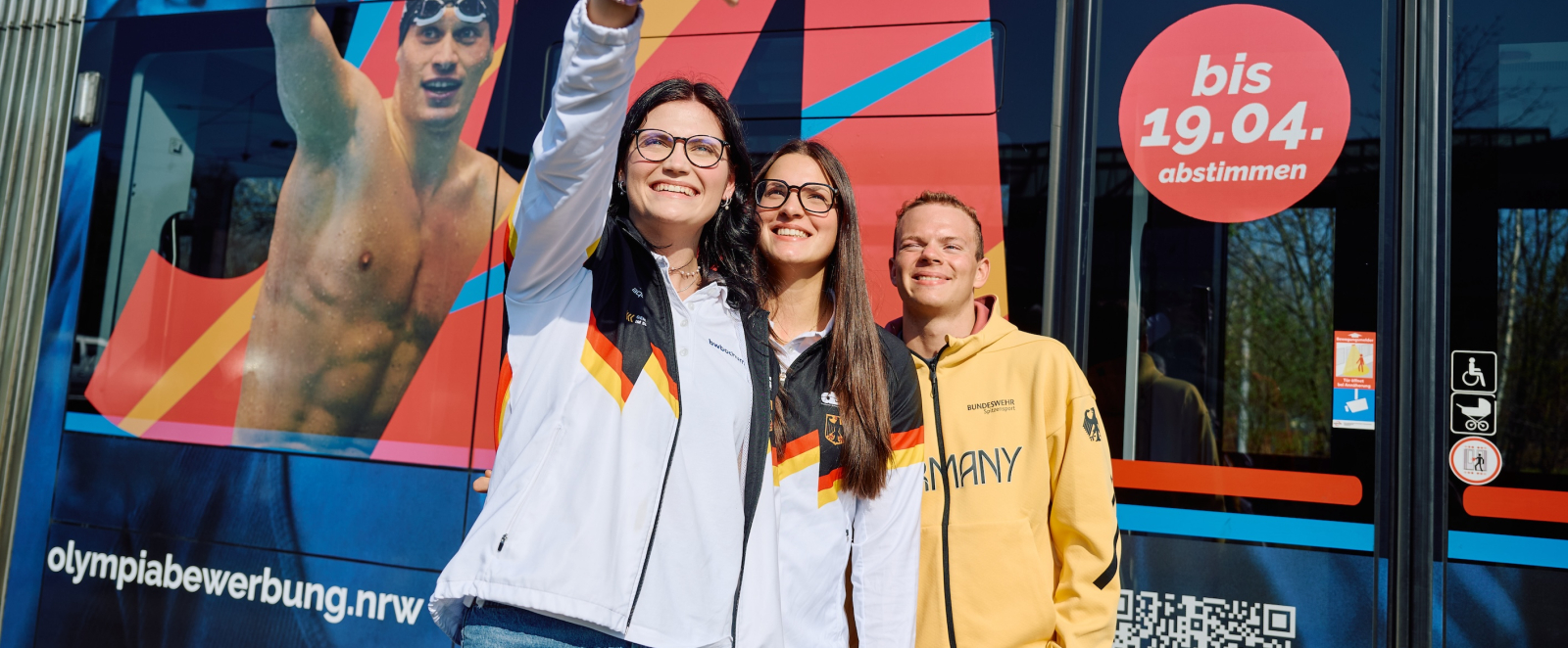 Zwei junge Frauen in hellem Oberteil sowie ein junger Mann im gelben Team D-Hoodie vor einer beklebten Straßenbahn