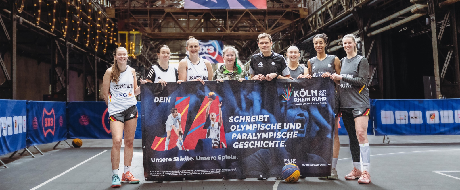 Andrea Milz mit 3x3-Aktiven und weiteren Personen in einer Sporthalle, sie halten ein Banner für die Bewerbung KölnRheinRuhr