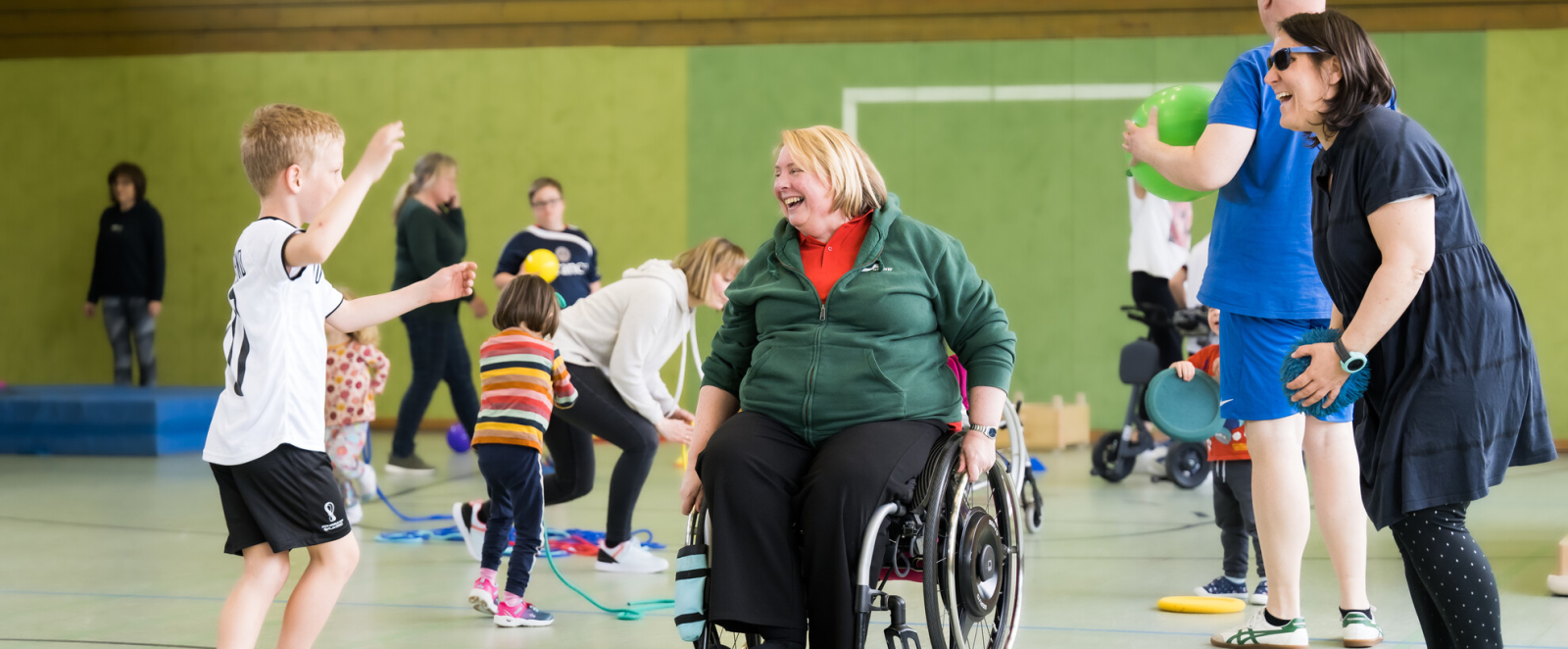 Ältere Frau in Sportkleidung in einem Rollstuhl ist umgeben von jüngeren Sportleren in einer Sporthalle