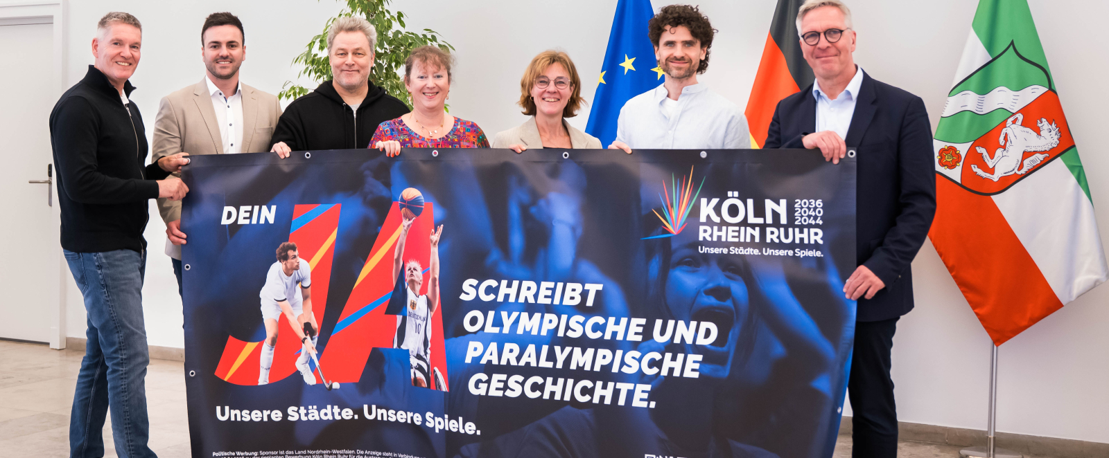 Sieben Personen, darunter Andrea Milz, halten ein Banner der Bewerbung KölnRheinRuhr