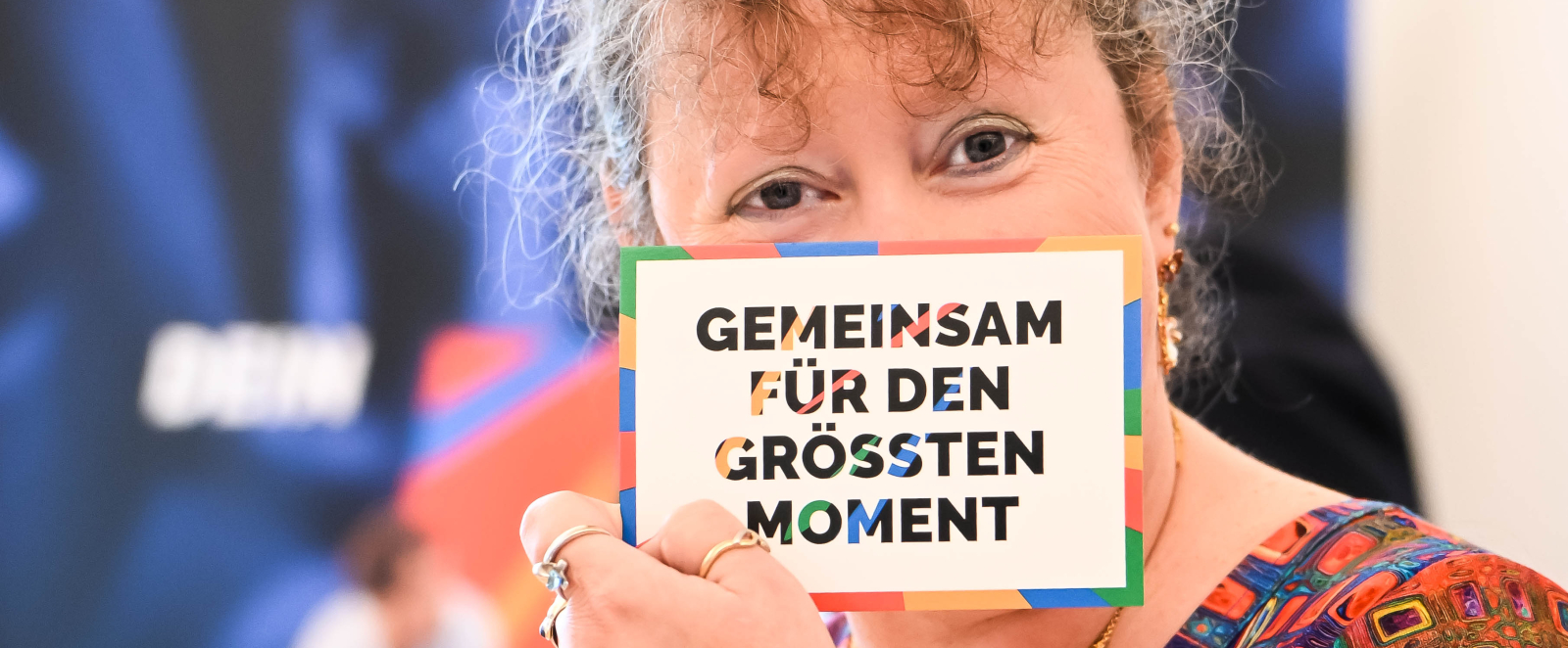 Andrea Milz hält ein Schild "Gemeinsam für den großen Moment" vor ihr Gesicht