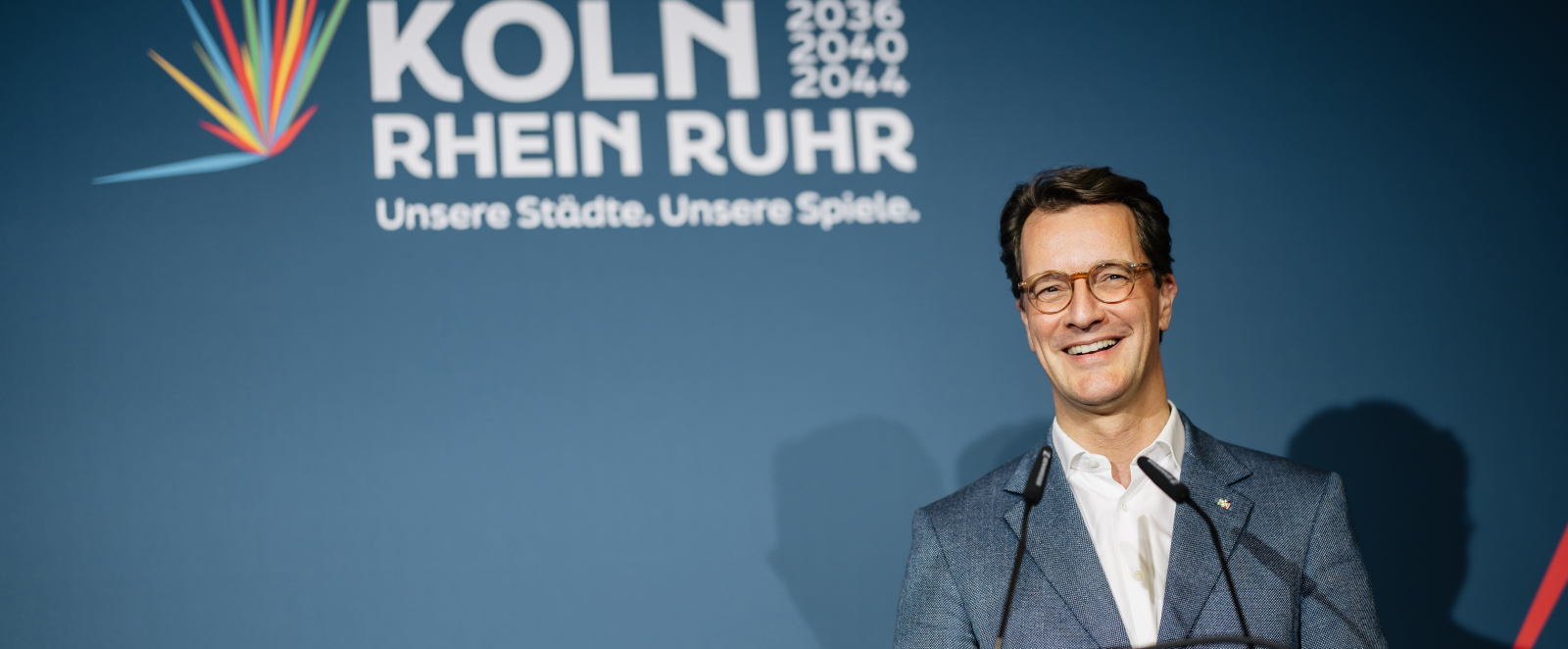 Ministerpräsident Wüst an einem Rednerpult vor blauem Hintergrund mit dem Logo der Bewerbung KölnRheinRuhr