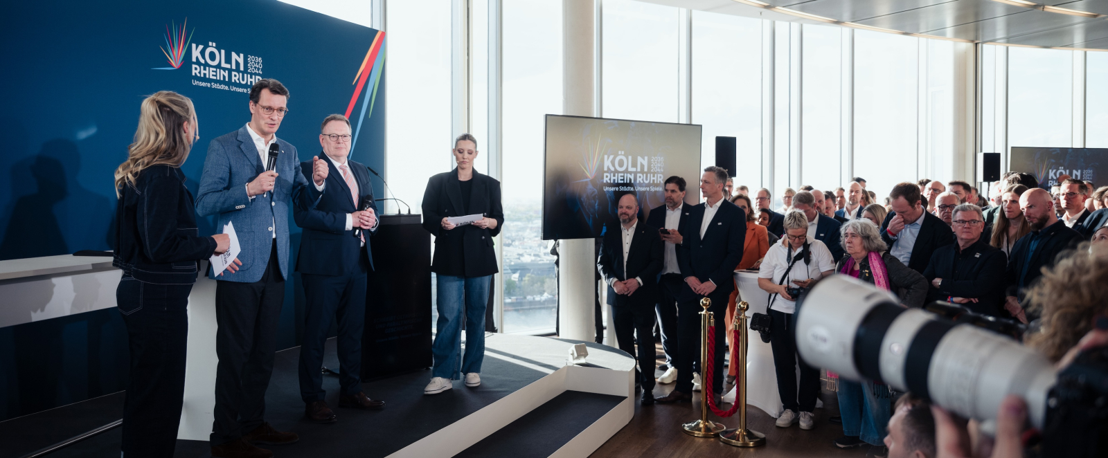 Ministerpräsident Wüst mit weiteren Personen auf der Bühne in einem Raum mit vielen hohen Fenstern