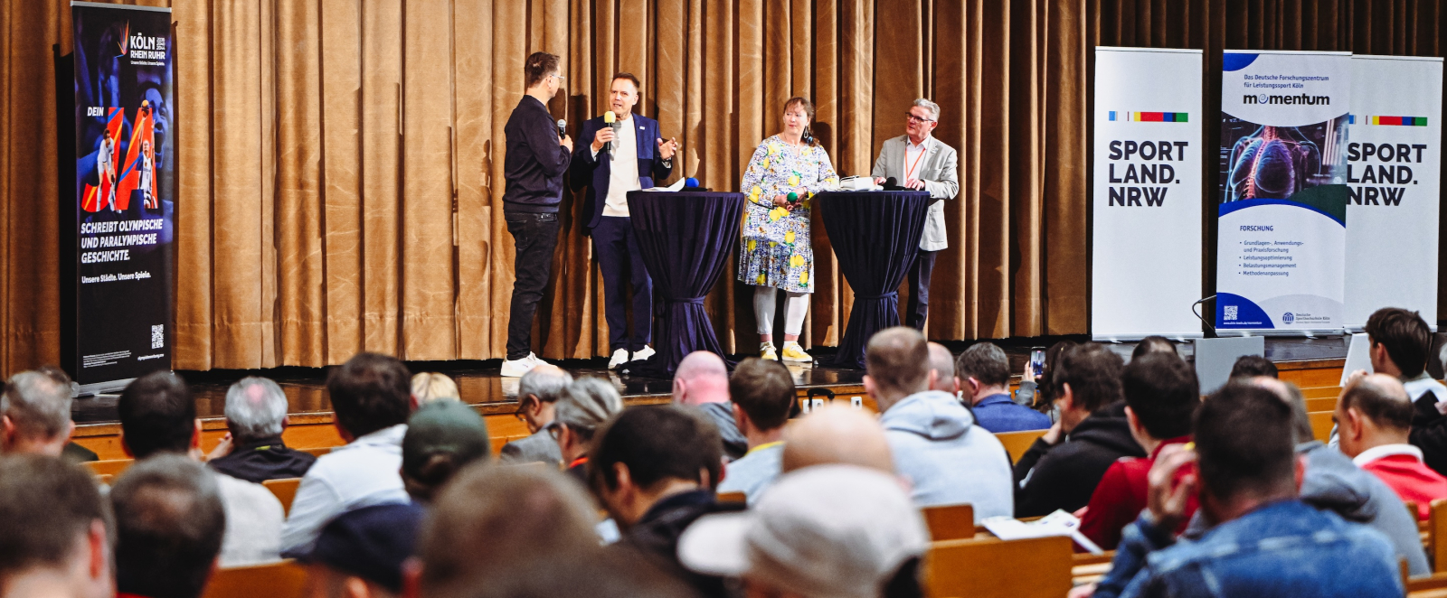 Personen auf einer Bühne mit einem Vorhang im Hintergrund, Personen auf Stühlen sitzen davor