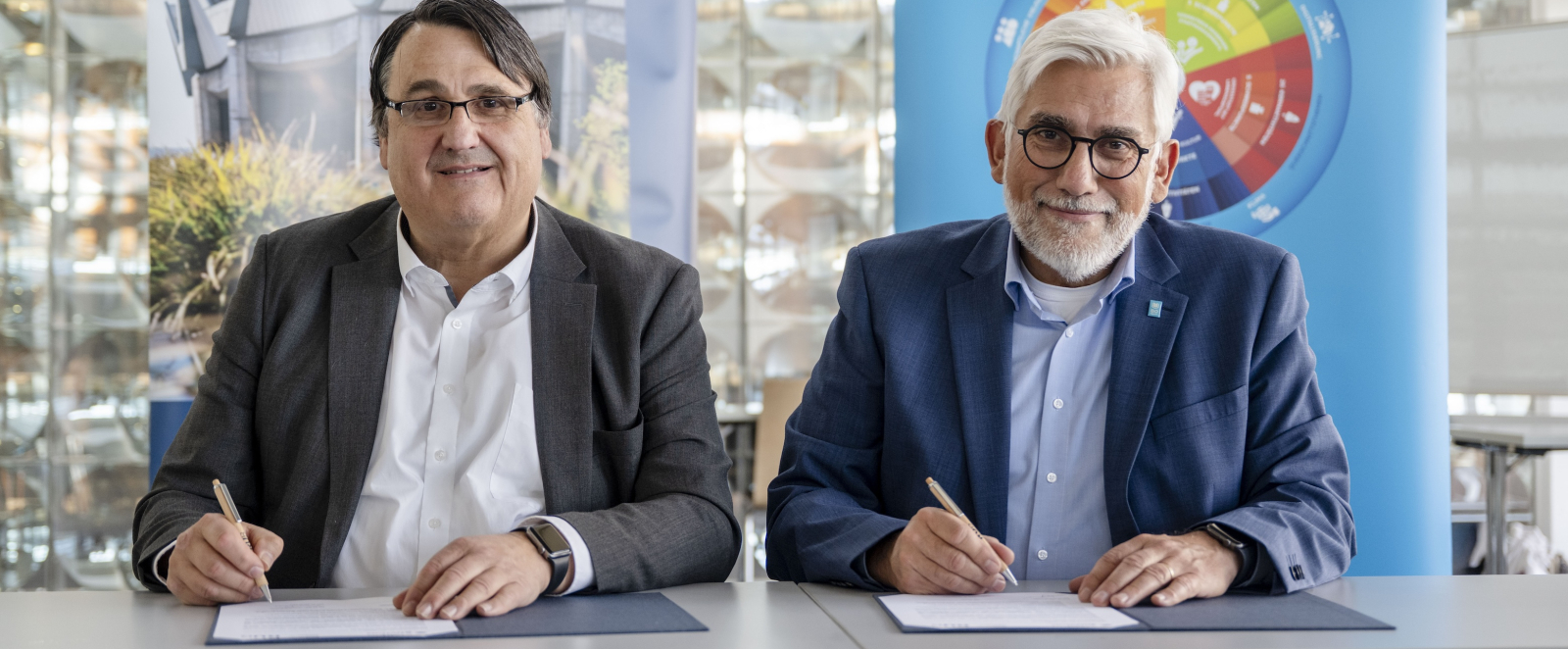 Zwei Männer an einem Tisch vor zwei bunten Bannern und einer Wand aus Glasbausteinen unterschreiben Dokumente