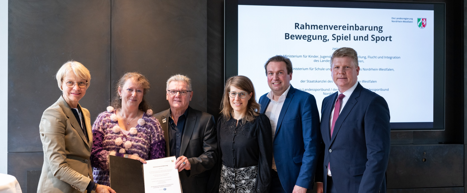 Sehcs Personen vor einem Screen mit der Aufschrift "Rahmenvereinbarung Bewegung, Spiel und Sport"