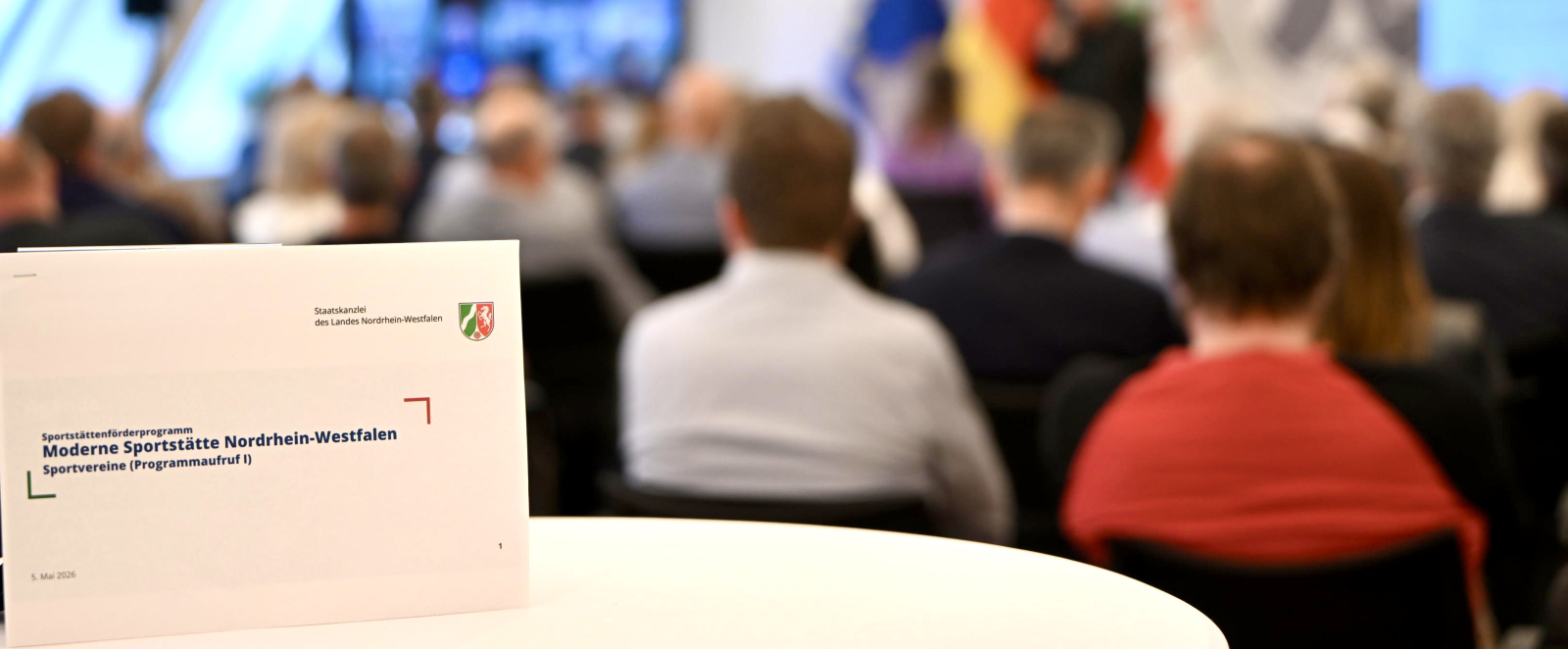 Schild mit "Moderne Sportstätte" auf einem Tisch, man sieht Teilnehmende einer Pressekonferenz von hinten