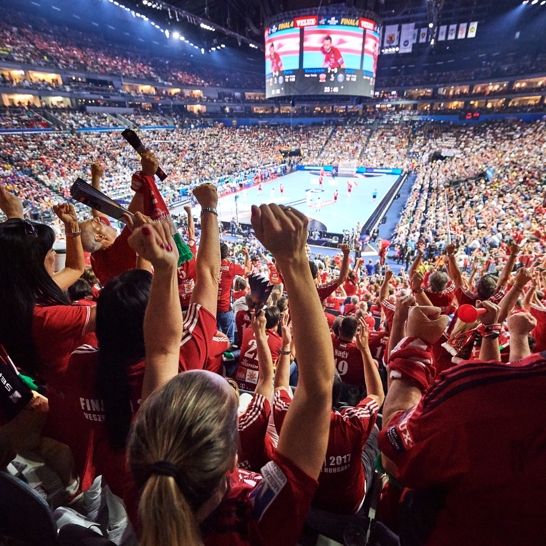 Man sieht von der hinteren Reihe in dievollbesetzte, geschlossene Mehrzweckhalle Lanxess Arena. Viele Zuschauer haben den rechten Arm oben und jubeln ihrer Mannschaft zu. Oben in der Mitte hängt eine kreisrunde Bildschirmpräsentator an der Decke.