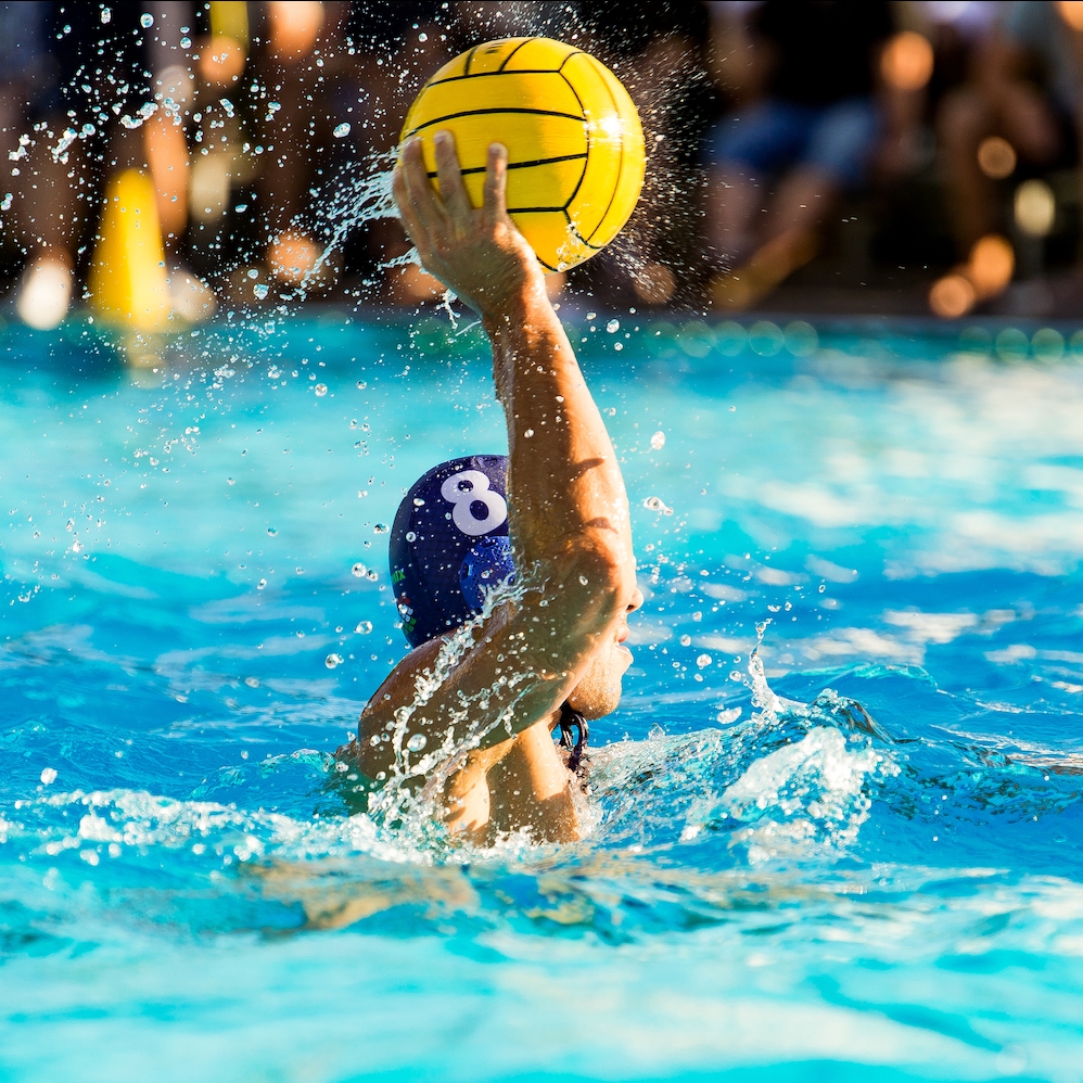 Ein Mann mit blauer Badekappe in einem Schwimmbecken hält einen gelben Wasserball in der Hand und holt zum Wurf.