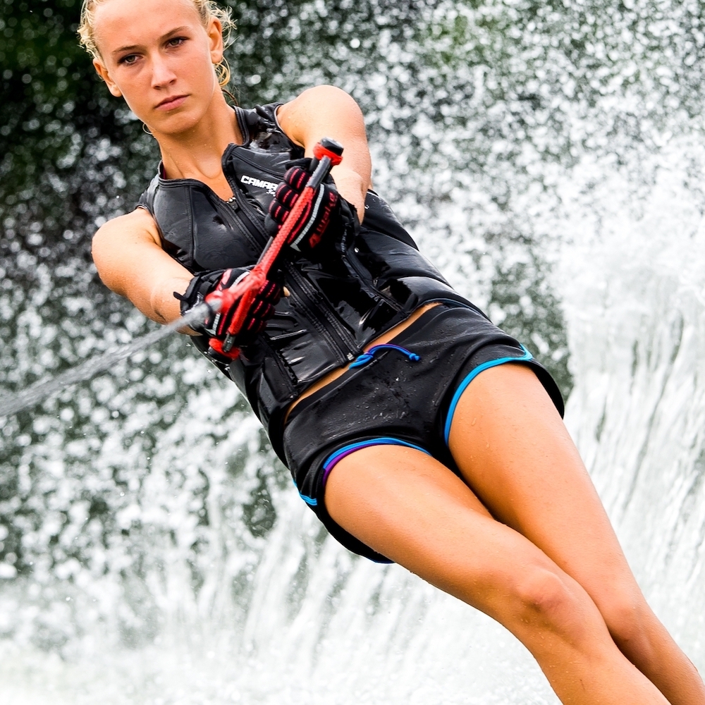 Das Bild zeigt eine schlanke, blonde Frau in schwarzer Wassersportbekleidung und schwarzer Schwimmweste beim Wasserski fahren.