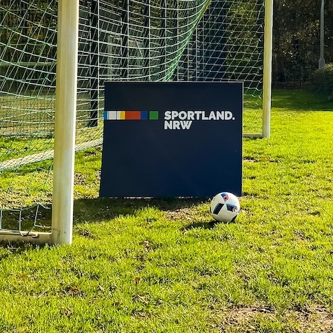 Sportland NRW Schild mit einem Ball auf einer grünen Fußball Wiese, hinter dem Schild ist ein Fußballtor