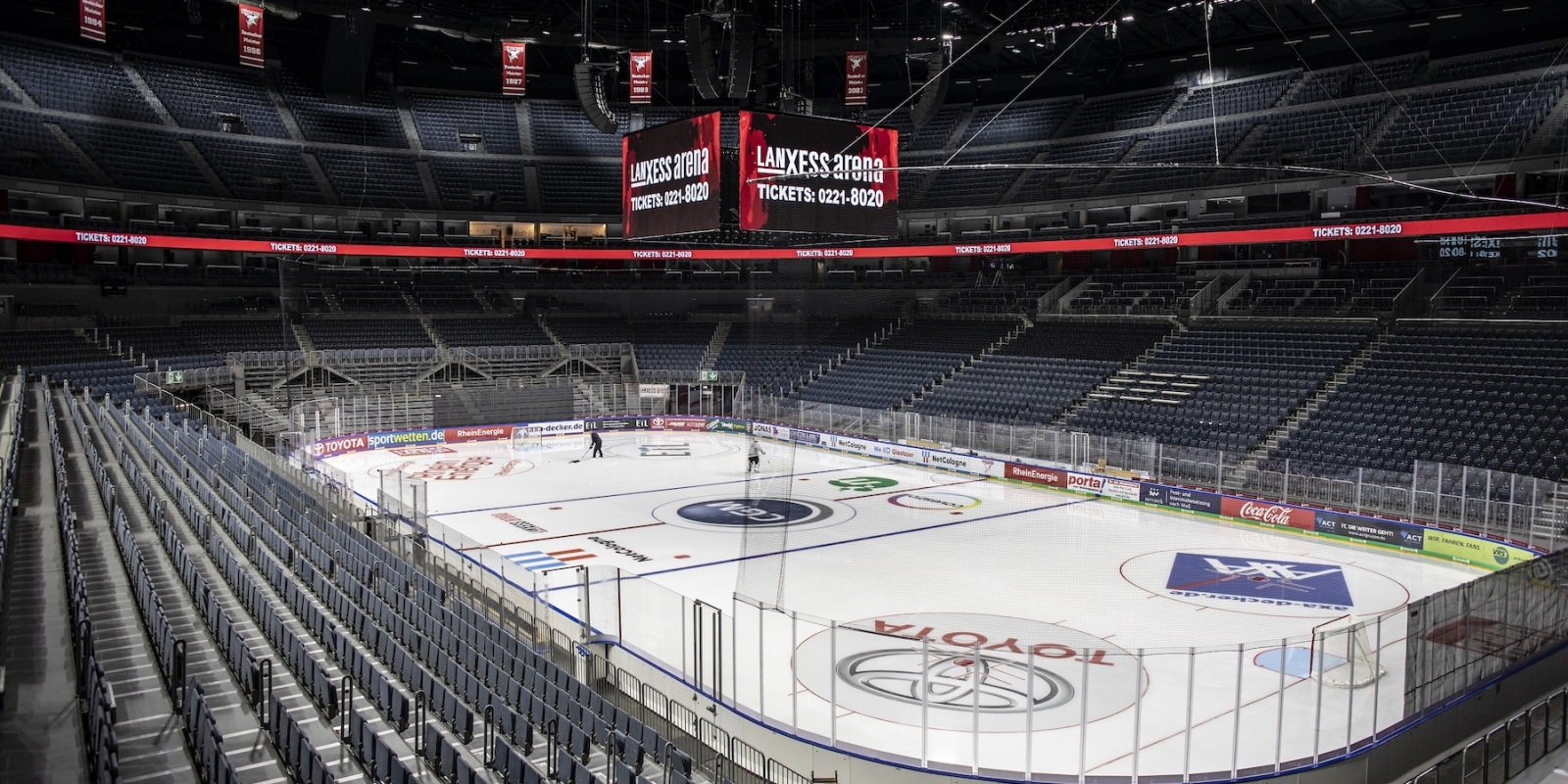 Sporthalle LANXESS Arena in Köln aus der Innenansicht, oben an der Decke hängt ein vierseitiger Bildschirmpräsentator. Die Tribühnen sind leer, auf der Eisfläche des Eishockeyfeldes trainieren zwei Eishockeyspieler.