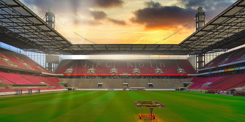 RheinEnergieSTADION Köln aus der Innenansicht. Zu sehen ist der grüne Rasen und die roten Stadionsitze mit Schriftzug 1.FC Köln