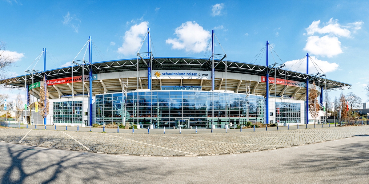 Das Stadion schauinsland-reisen-arena Duisburg aus der Außenansicht. Es hat im Eingagnsbereich ein breite und hohe Glasfront, die Außenstützpfeiler sind royalblau gefärbt.