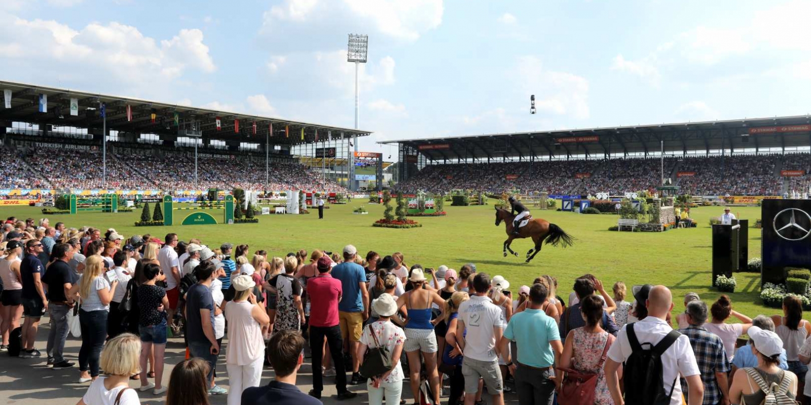 Zuschauer betrachten einen Reiter, der auf seinem Pferd im Aachener CHIO Stadion auf ein Hindernis zu gallopiert. 
