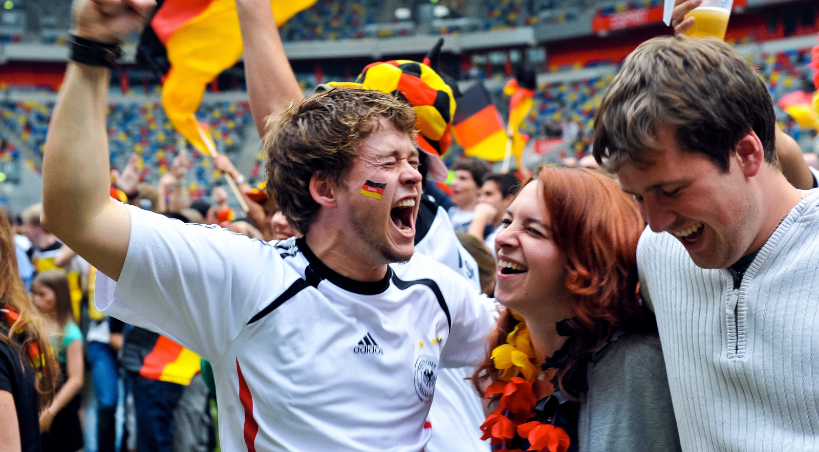 Jubelnde Fans mit Deutschland-Flaggen i einem Stadion. Im Vordergrund ein Fan in weiß-schwarzem Trickot und Deutschland-Flagge auf der Wange.