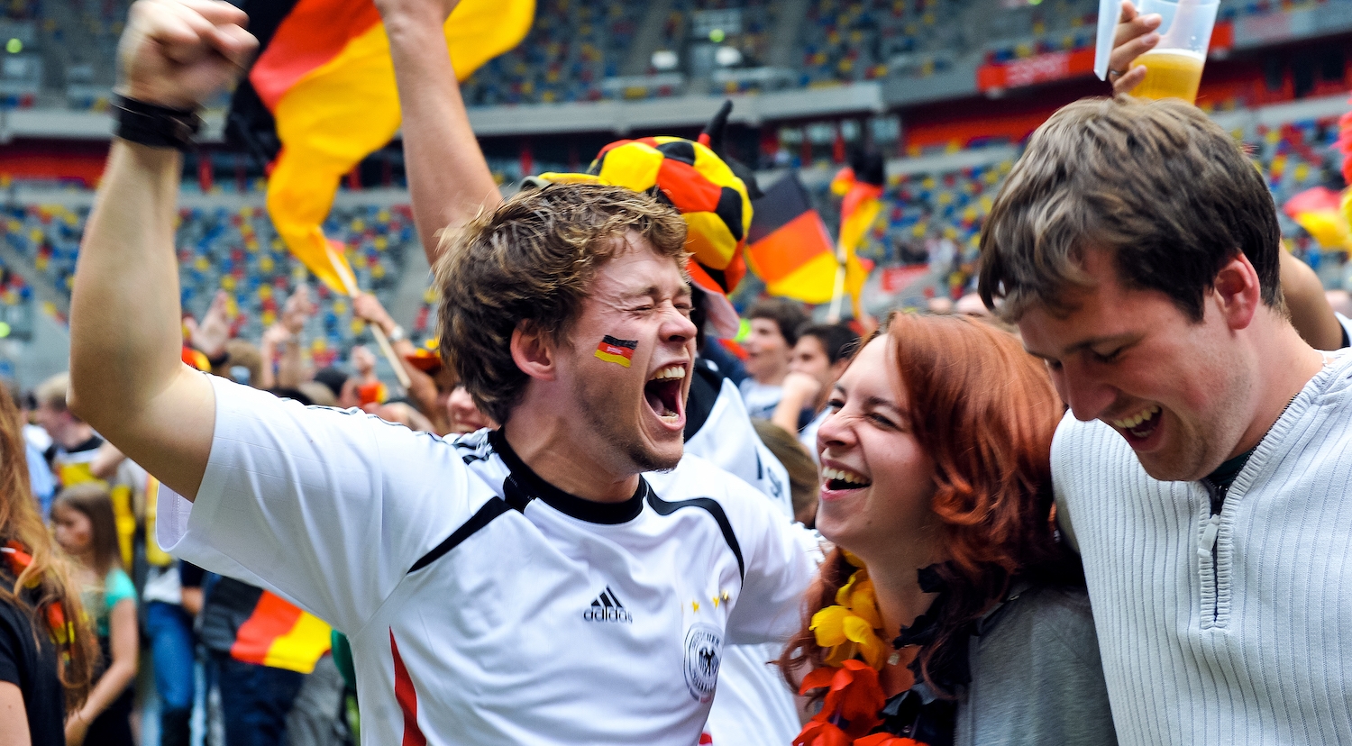 Jubelnde Fans mit Deutschland-Flaggen i einem Stadion. Im Vordergrund ein Fan in weiß-schwarzem Trickot und Deutschland-Flagge auf der Wange.