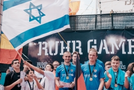 Viele Sportler stehen auf einer Bühne, einige Sportler halten Flaggen. Man sieht die Flaggen von Deutschland und Israel. Auf der Rückwand steht weiß auf schwarzem Grund "Ruhr Games".
