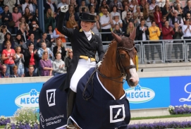 Pferdesport - Eine Reiterin auf einem brauenen Pferd mit dunkelblauen Pferdeumhang auf dem weiß gedruckt steht "CHIO Aachen".