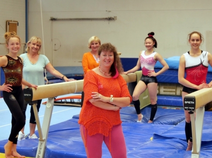 Staatssekretärin Milz in orangefarbenem T-Shirt und Sporthose mit sechs Damen in einer Turnhalle. Zwischen den Damen stehen zwei Schwebebalken, auf dem Boden liegen blaue Bodenmatten.