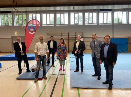 Staatssekretärin Andrea Milz steht mit mehreren Vereinsmitgliedern in einer Turnhalle, überall sidn verstreut Matten auf dem Boden verteilt.