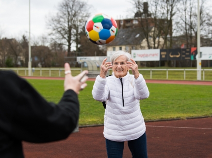 Frau Erika Rischko in weißer Daunenjacke und schwarzer Hose wirft der Staatsekretärin Andrea Milz in schwarzen Sportbekleidung einen bunten Sportland.NRW-Fußball zu. Die sechseckigen Felder des Fußballs haben die Sportland.NRW-Farben Weiß, Rot, Gelb, Grün, Blau und Dunkelblau.