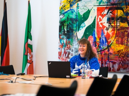 Staatssekretärin Milz sitzt an einem beigen Besprechungstisch mit schwarzen Stühlen. Vor sich ein schwarzes Notebook, links von ihr die Flaggen von Detuschland und NRW. Hinter sich an der Wand befindet sich ein großes, farbiges Kunstwerk mit einem großen NRW-Wappen oben mittig, verschiedene farbige Motive wie der Kölner Dom, die Hohenzollernbrücke gehen farblich abwechselnd ineinander über.