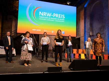 Staatssekretärin Milz steht mit mehreren Persoenen auf einer Bühne. HInter ihnen das bunte Logo vom NRW-Preis mit einem regenbogenfarbigen Farbverlauv von unten nach oben, von rot über orange, gelb, grün, hellblau, blau bis zu viiolett.