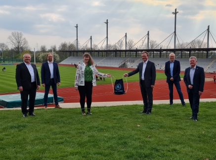 Staatssekretärin Andrea Milz steht mit 5 weiteren Menschen vor einer Sportstätte mit Tartanbahnen und einem Rasenplatz in der Mitte.