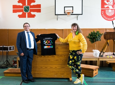 Staatssekretärin Milz hält ein schwarzes T-Shirt mit Logo der Förderung mit der Zahl 500 in Regenbogenfarben.