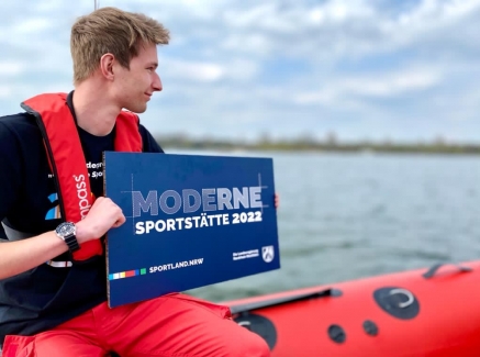 Ein junger Mann sitzt auf einem Schlauchboot und hält ein blaues Schild 