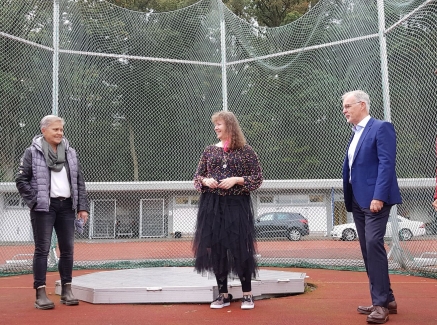 Staatssekretärin Andrea Milz steht mit mehreren Vereinsmitgliedern auf einem Hammerwurf-Sportplatz. Im Hintergrund sieht man das Auffang-Stahlnetz im Halbkreis aufgestellt.