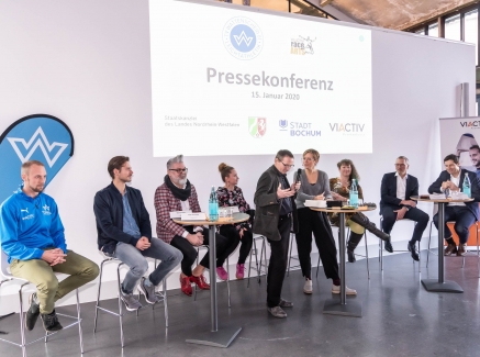 Staatssekretärin Andrea Milz sitzt mit Teilnehmern der Pressekonferenz auf Hockern an Rundtischen. Hinter ihnen eine weiße Rückwand mit einer Präsentation.