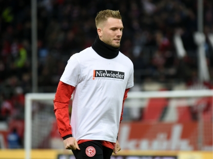 Ein Fußballer mit weiß-rotem FC-Trikot und schwarzer Hose geht über den Sportplatz, auf seinem T-Shirt steht Weiß auf Schwarz "Nie Wieder".