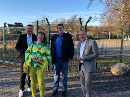Staatssekretärin Milz in gelb-grünem Strickpullover und gelbgrüner Hose steht mit drei Herren vor einem grünen Zaun.