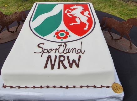 Sportland.NRW-Kuchen, im Hintergrund drei Reiter auf Pferden