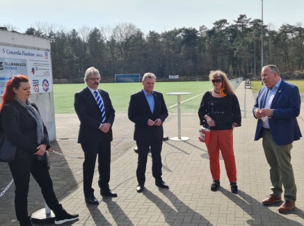 Staatssekretärin Milz steht mit mehreren Personen im Halbkreis vor einem Rasensportplatz.