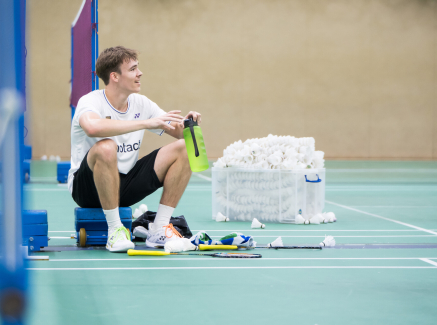 Linus sitzt mit angezogenen Knien in der Badmintonhalle am Netz