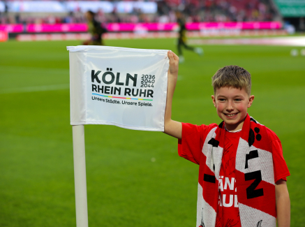 Junge in rotem Trikot auf dem Rasen im RheinEnergiestadion hält die Fahne "KölnRheinRuhr"