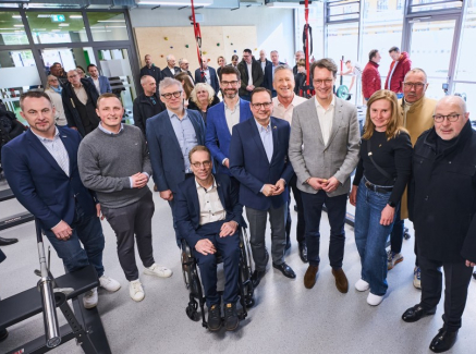 Hendrick Wüst inmitten mehrerer Personen, davon einer im Rollstuhl, in einem Raum mit Fenstern im Hintergrund