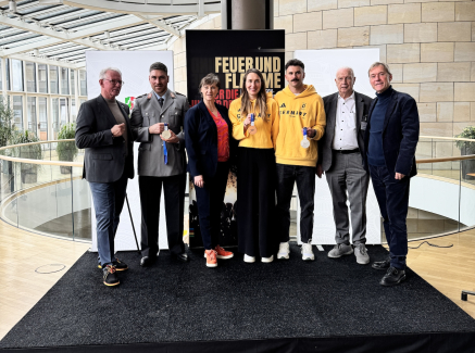 Sieben Personen auf einem schwarzen Podest im Landtag NRW mit vielen Fenstern im Hintergrund