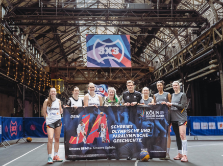 Andrea Milz mit 3x3-Aktiven und weiteren Personen in einer Sporhalle, sie halten ein Banner für die Bewerbung KölnRheinRuhr