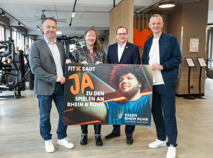 Andrea Milz mit drei Männern, im Hintergrund ist ein Fitnessstudio zu sehen, sie halten ein Banner "Fitx sagt Ja zu Spielen an Rhein & Ruhr"