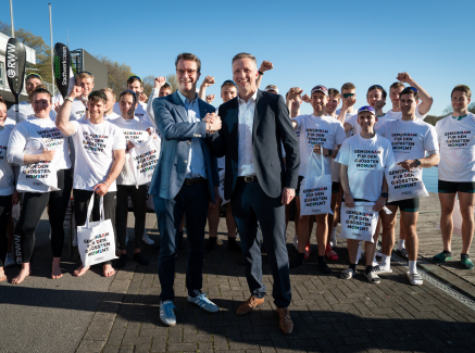 Hendrik Wüst umgeben von Ruderen in weißen Shirts mit der Aufschrift "Gemeinsam für den Größten Moment"